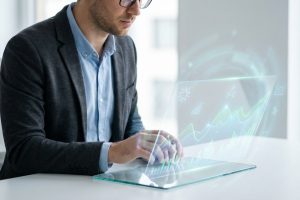 Un homme aux lunettes travaille sur un écran transparent interactif affichant des graphiques et données lumineux dans un bureau moderne.