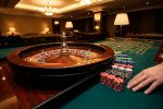 Main d'un joueur, jetons et roue de roulette en bois sur une table verte dans un casino luxueux avec des lumières chaudes.